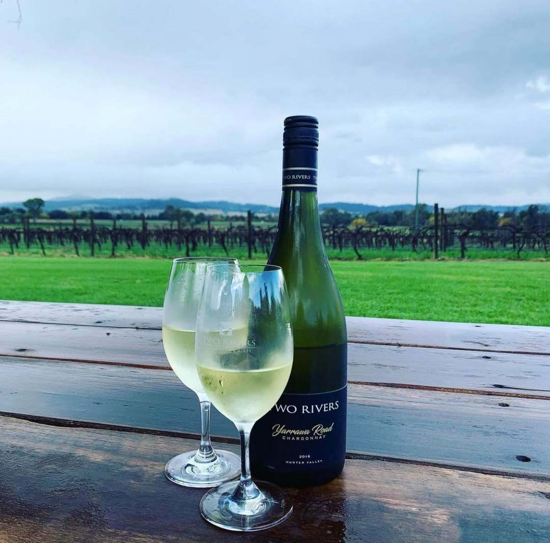 A glass of Semillon and the bottle on a timber table, vines behind